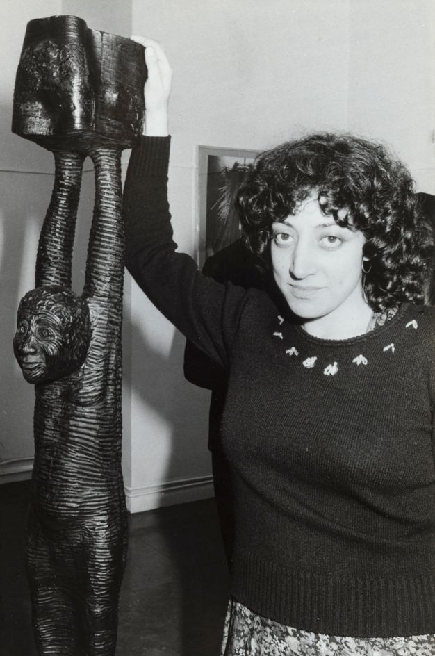 Black and white photograph. A woman with black curly hair falling her shoulders stands next a sculpture as she looks at us. The sculpture is of a person with their arms raised. She rests one hand on the top, which comes up over her head.