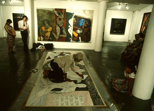 Installation. A person draws on a work of art spread on the floor at the centre of a gallery hung with paintings. The artist has brown skin, white hair, and is barefoot.
