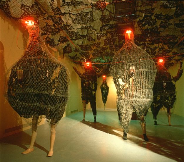 Installation. Five sculptural forms stand in a long gallery space. Each has the legs of a mannequin and bodies that are either humanoid or shaped more like a chicken. Each body has black-coloured material encased in chicken wire, and red lights glow like eyes from the very tops of tiny heads.