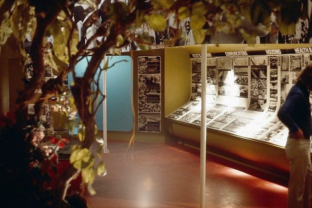 Installation. Inside a gallery, we look through tree branches at a display of black and white photographs that covers the far wall and curves down and towards us, making a low, angled counter. The headings over the images that we can make out read “Communications”, “Housing”, “Health &”, with the final one cut off.