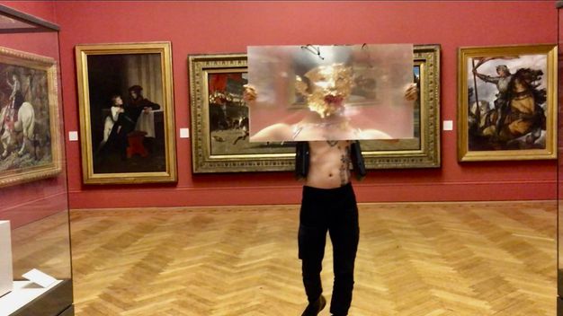 Performance still. A medium-skinned holds a sheet of magnifying plastic, arms stretched wide, as he walks towards us a gallery hung with paintings in gold frames. His face is covered in gold leaf, and his mouth is filled with red chili peppers.