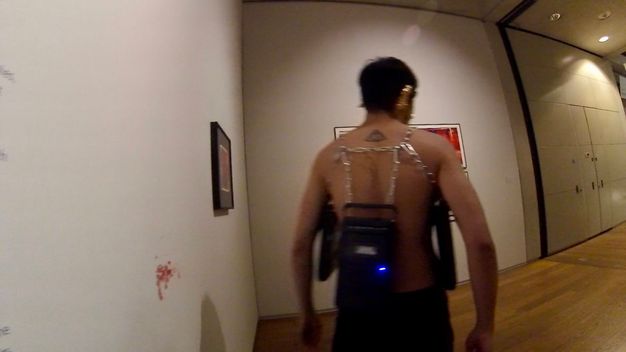 Performance still. A medium-skinned, bare-chested man walks away from a bloody mark on a gallery wall. Cassette players hang from chains around his torso.