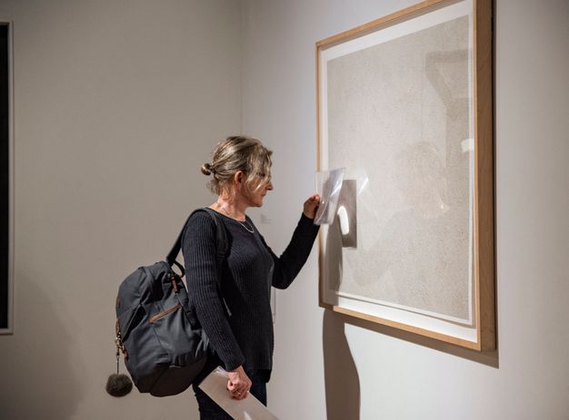 A woman holds a sheet of magnifying material, about the size of a tablet, up to a framed, white work of art, looking closely at the surface.