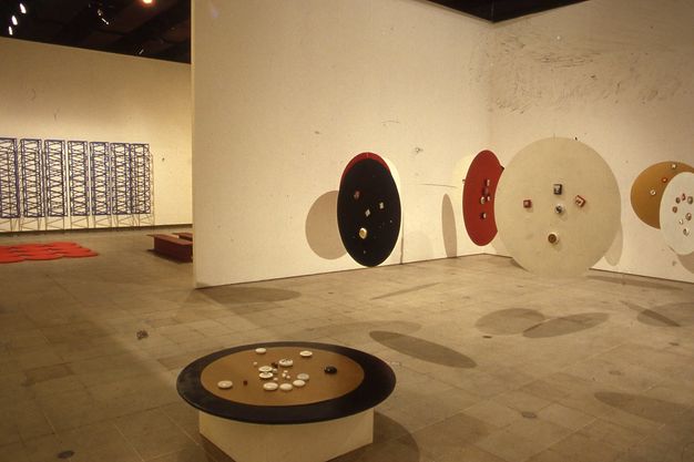 Installation. At least eight round discs, like tabletops, float suspended from a gallery ceiling. A seventh sits on a pedestal and holds round stones or objects made of another material. The hanging discs are painted brown, red, or black. A sculptural piece hanging on the wall seen through a doorway to our left is of ladder or trellis-like structures creating a dense geometric pattern.