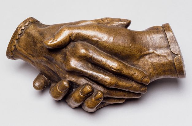 Sculpture. Bronze cast of two right hands holding each other. The hands cut off at the wrists, and one is edged in a straight cuff and the other in scalloped cuff. In this view, one rests along the palm of the other. Veins on the back of one hand and on the underside of the wrist of the other are visible. The patina is honey brown.