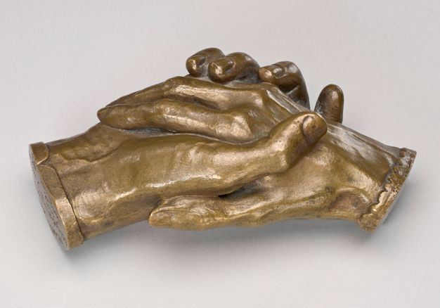 Sculpture. Bronze cast of two right hands holding each other. The hands cut off at the wrists, and one is edged in a straight cuff and the other in scalloped cuff. In this view, the thumbs tuck in against each other. Veins on the back of one hand and on the underside of the wrist of the other are visible. The patina is an almost flat gold colour.