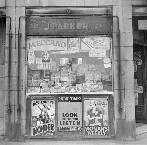 Black and white photograph. A storefront is packed with publications and ads for cigarettes and candy bars. One sign reads, “Fountain Pens Repaired Quick Service”. Three posters are affixed to the wall under the window.