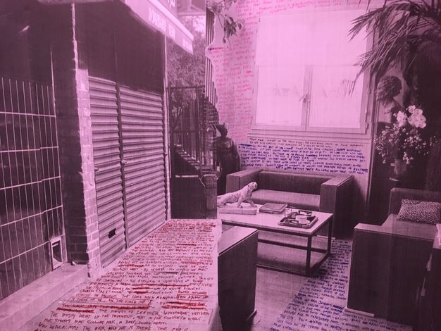 Installation. What initially looks like a single black and white photograph tinted magenta is two views, one of a shuttered storefront and one of the inside of a room with couches and a coffee table. Text in red, blue, or pink appears to have been written on the pavement in front of the store and the floor and walls of the room.