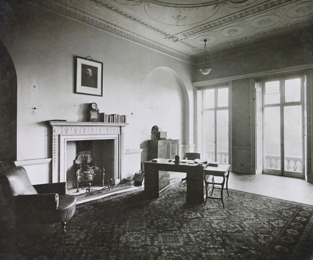 Black and white photograph. A high-ceiling room is furnished sparsely with a patterned rug, a small desk and table, a cabinet, and an armchair. About a dozen books are lined up on the mantle below a portrait hanging above. The mantle and ceiling are decorated with plasterwork, and windows across from us span nearly the height of the walls.