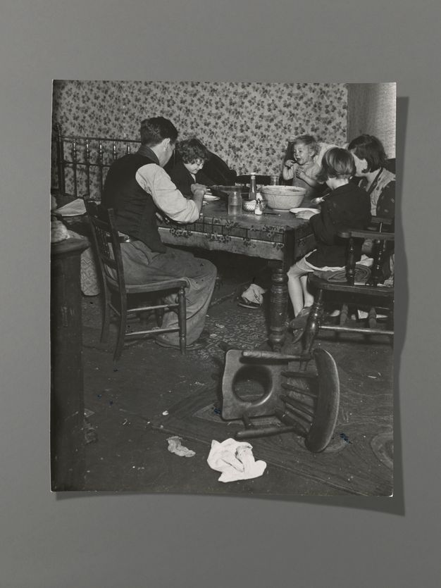 Black and white photograph. A man, woman, two children, and a baby gather around a dining table. Most have their backs to us, but one child and the baby face us, smiling. An overturned commode chair is between us and the group.