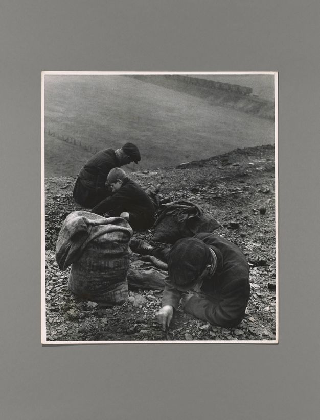 Black and white photograph. Two men and a boy sit or lean against a steep incline, picking through dirt and pebbles. A sack sits next to the man closest to us. A flat field beyond the incline leads back to a train in the distance.