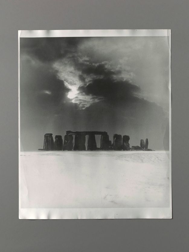 Black and white photograph. The dark posts and lintels of Stonehenge mark the transition from a stark, snowy field to a bright sunburst piercing dark clouds above.
