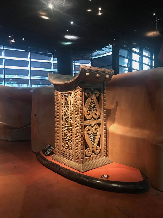 A rounded seat is atop an elaborately pierced and carved rectangular base. The set curves like a shallow letter C. The object is spotlit in a museum with windows lining the far wall.