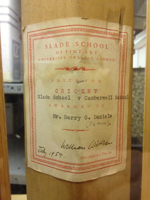 Cricket bat. A label printed with a red border and text is affixed to the bat. It begins, “Slade School of Fine Art University College London”. Below, red text, black typewritten letters, and a handwritten signature and date fill the rest of the label.