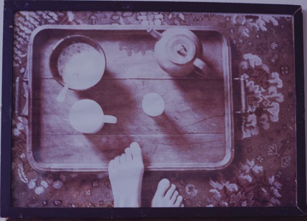Colour photograph. In tones of violet and smoky purple, we look down onto a wooden tray holding a teapot, mug, and two more vessels, one with a spoon. The carpet beneath is woven with a stylized, floral design. The toes of one foot creep up over the bottom edge of the tray, and the other foot stands next to it. The photograph is in a purple frame.