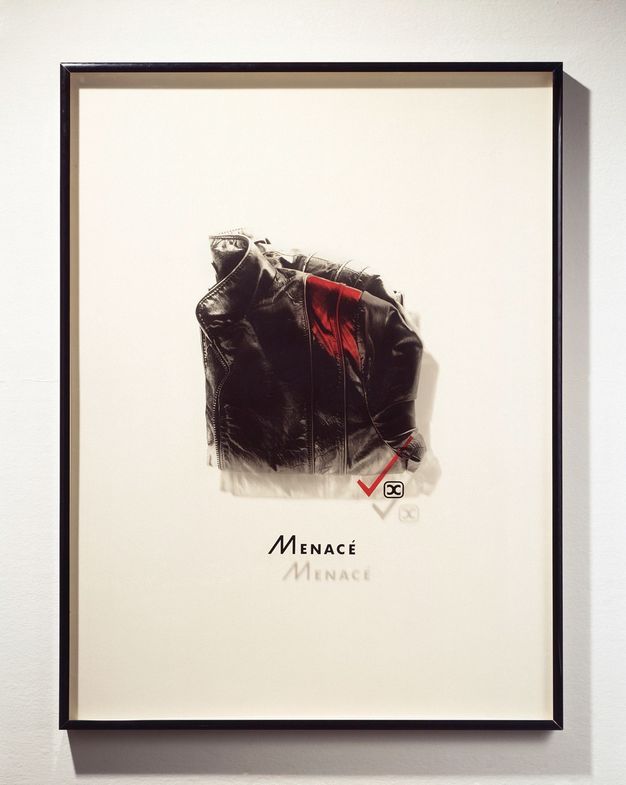 Mixed media. A photograph of a high-collared, folded, black leather jacket has been printed onto a piece of Plexiglass and mounted in a frame so it casts a shadow on the mat behind it. A swipe of red has been added to the front shoulder of the jacket. The word “Menacé” is below.