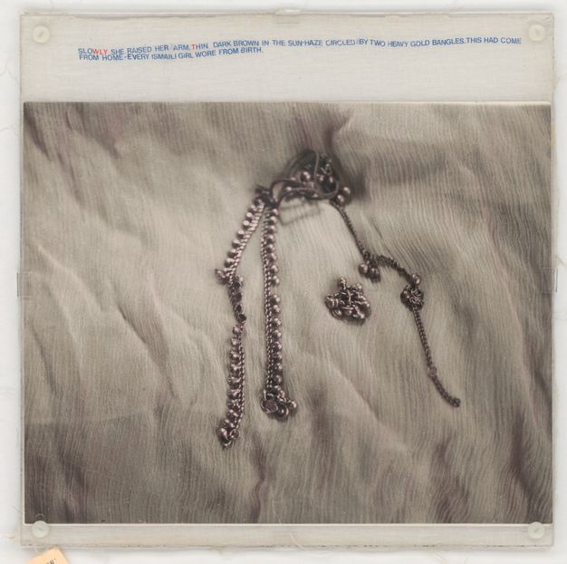 Mixed media. A black and white photograph shows a piece of silver jewellery lying on a white, wrinkled cloth. Mostly blue text in all caps printed on muslin above the image reads, “Slowly she raised her arm, thin dark brown in the sun-haze circled by two heavy gold bangles. This had come from home – every Ismaili girl wore from birth”. The WLY in “slowly” and T in “thin” are red.