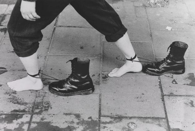 Film still. Shown from the hips down, a barefooted person walks on a sidewalk in this black and white image. One foot is planted far in front of the other, and each is tied around the ankle with the long laces of distinctive high, black Doc Martens boots.