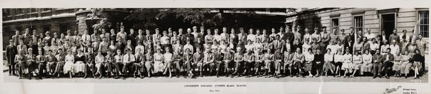 Black and white photograph. Around 125 men and women stand or sit with arms crossed, by their sides, or in their laps in four long lines in this panoramic class photograph. Most have pale skin, but there are a few men with darker skin. The men mostly wear suits and ties, the women dresses.