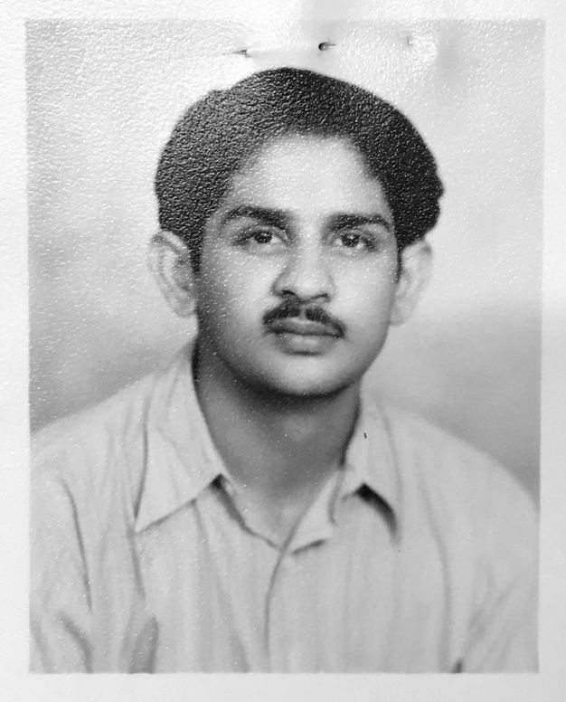 Black and white photograph. Shown from the shoulders up, a man looks at us. He has dark hair and brows, a wide nose, and a moustache over full lips. Light skips across the textured surface of the photo paper.