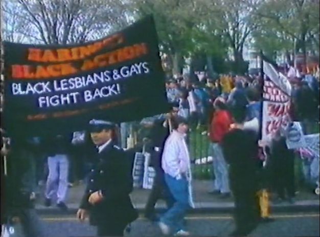 Video still. People gather, some walking, around a black banner with red and white text. Red text in all caps at the top reads “Haringey Black Action”. Below, white text reads, “Black Lesbians & Gays Fight Back!” A police officer stands between us and the crowd, which spill into a park or grassy area across the street from us.