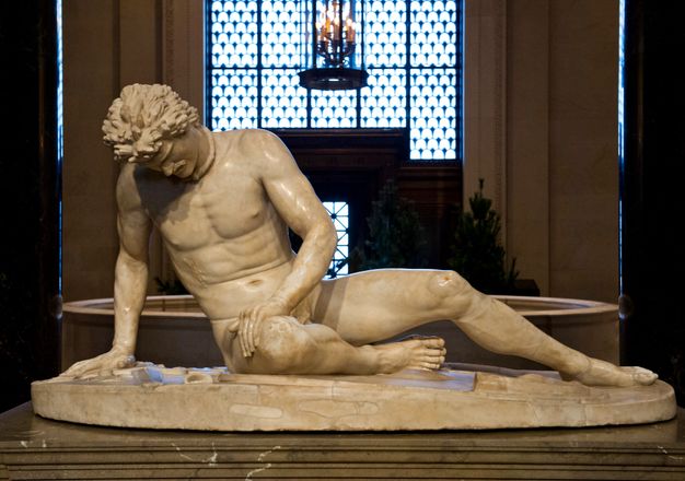 Sculpture. This freestanding statue shows a nude man sitting with his right leg tucked under him, the other extended. He leans on his right hand, and rests his other on his right thigh. He looks straight down so we see his furrowed brow, nose, and a hint of his moustache. A rings that opens at the front is around his neck, and blood dribbles out of a wound over his right ribs.