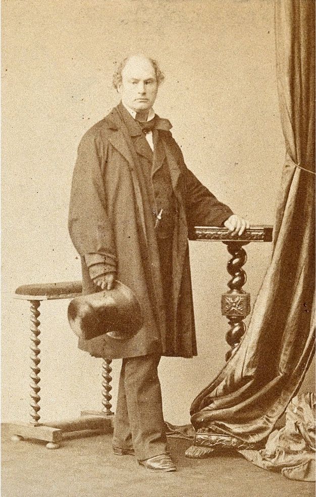 Black and white photograph. In warm tones of brown and parchment white, a pale-skinned, balding man poses in a studio. He is clean shaven and wears a baggy coat over a full suit. He holds a tall, shiny top hat in one hand, and the other rests on a tall stand with a table-top.