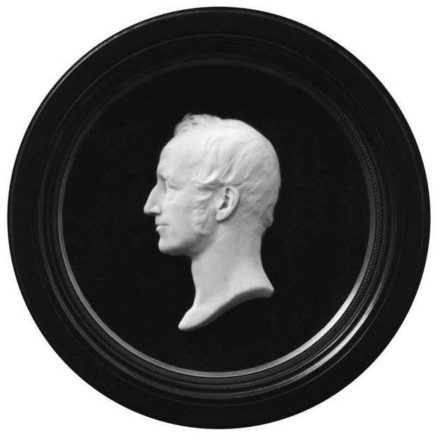 Cameo. A man’s profile facing our left is carved in white wax against a black background in a round medallion. He has a high hairline, bumped nose, and his thin lips nearly smile. His hair is swept forwards over his ears, and he has short mutton chops.