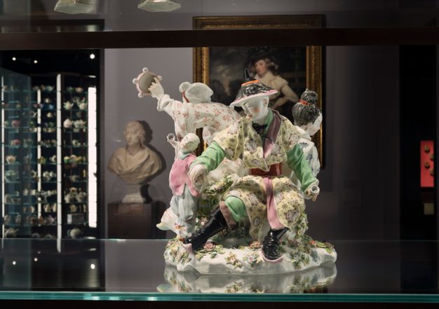 Porcelain. A grouping of at least three adults and one child are in a ring of activity and movement. One person holds a tambourine out, and the child holds up a small object as a man, closest to us, grips the child’s other hand. The porcelain is glazed with pastel yellow, spring green, rose pink, red, and black.