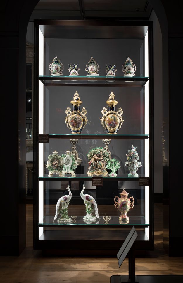 Installation. Four shelves are lined with glass on both long sides so we look through it. From the bottom up are a pair of birds on the bottom shelf and figural groups on the second shelf. Urns and vessels fill the rest of the space.