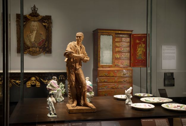 Installation. A case has four statuettes of people, one bird on a tree trunk, and five plates. A sideboard, chest of drawers, and a painting line the wall on the far side of the room.