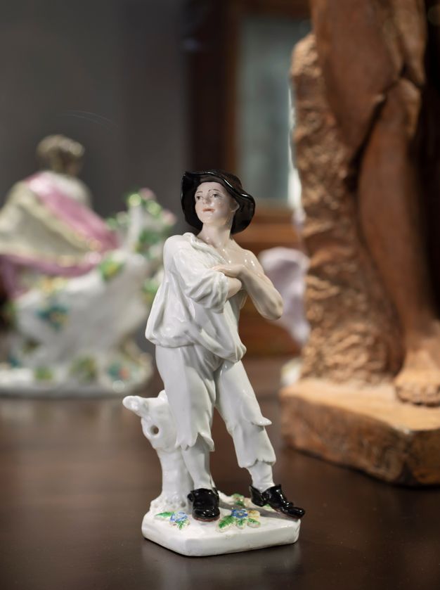 Porcelain. A figurine of a pale-skinned man stands with arms crossed, looking off to our left. He wears a wide-brimmed black hat, white, tattered clothes, and black shoes.