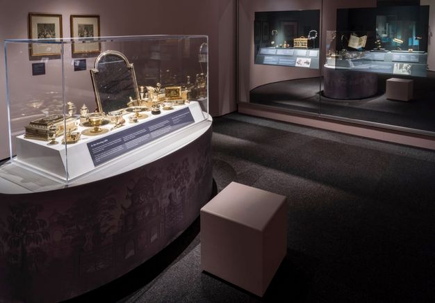 Installation. A long, oval-shaped platform holds a case filled with gold-coloured glass vessels, boxes, and a mirror.