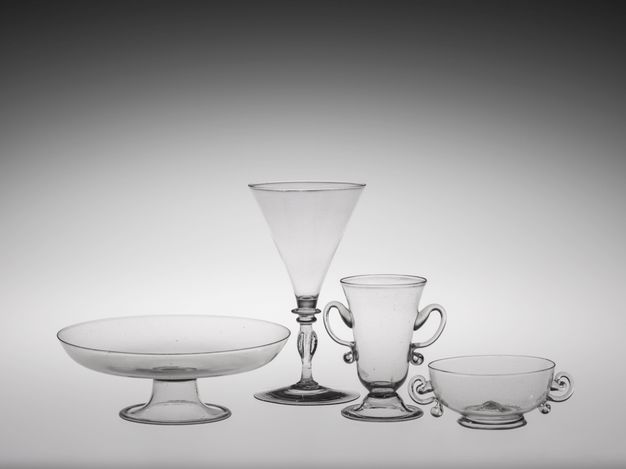 Glass. Two dishes and two glasses are clear blown glass. A tall posset glass and bowl have ear-shaped handles. The posset glass, a triangular wine glass, and a dish flare delicately at the lips.
