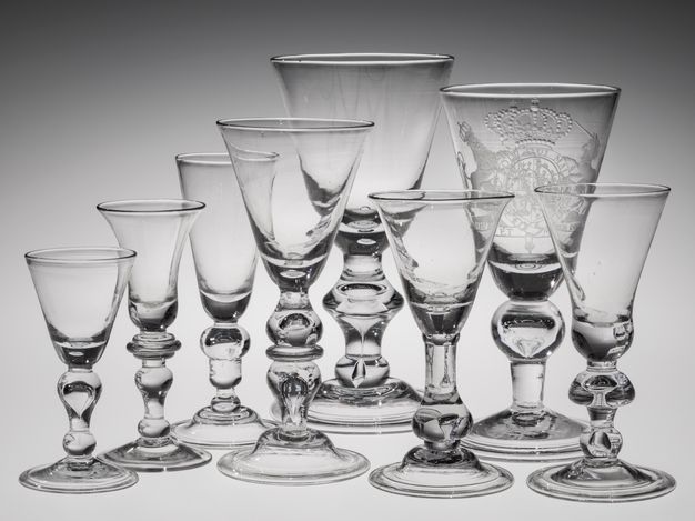Glass. Eight clear glass drinking vessels are shaped like upside-down triangles or bells. Each has a knob on a thick stem over a flaring foot. Most of the knobs are right under the bowl of the glass but one has a knob closer to the foot.