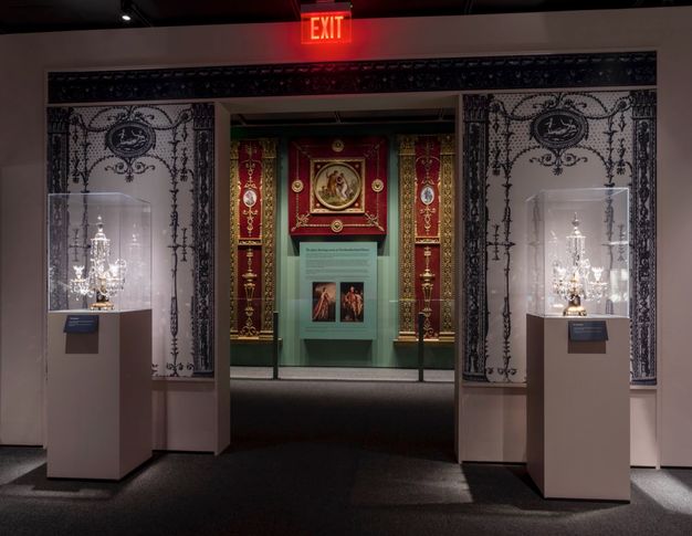 Installation. An opening in an exhibition space is flanked by windows that appear to be frosted and etched with designs reminiscent of wrought iron. Through the doorway is a set of three red panels with gold decoration; one is square and the other two reach from floor to ceiling.