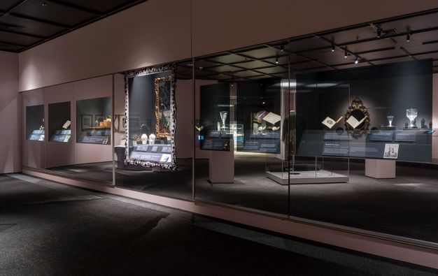 Installation. Display cases with glass, books, and other works of art are reflected in a long wall of mirrors. The space is dimly lit with spotlights on the cases and objects within.