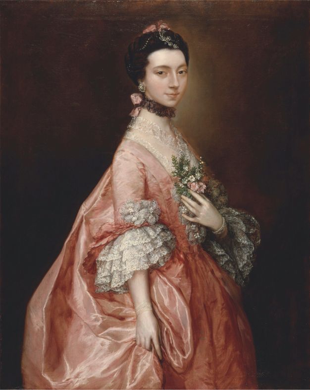 Painting. Shown from the thighs up, a pale-skinned woman wearing a carnation pink dress with cascading lace cuffs at the elbows stands in front of a dark background. Her body faces our right, her far hand resting on a spray of pink and white flowers and green leaves at her chest.