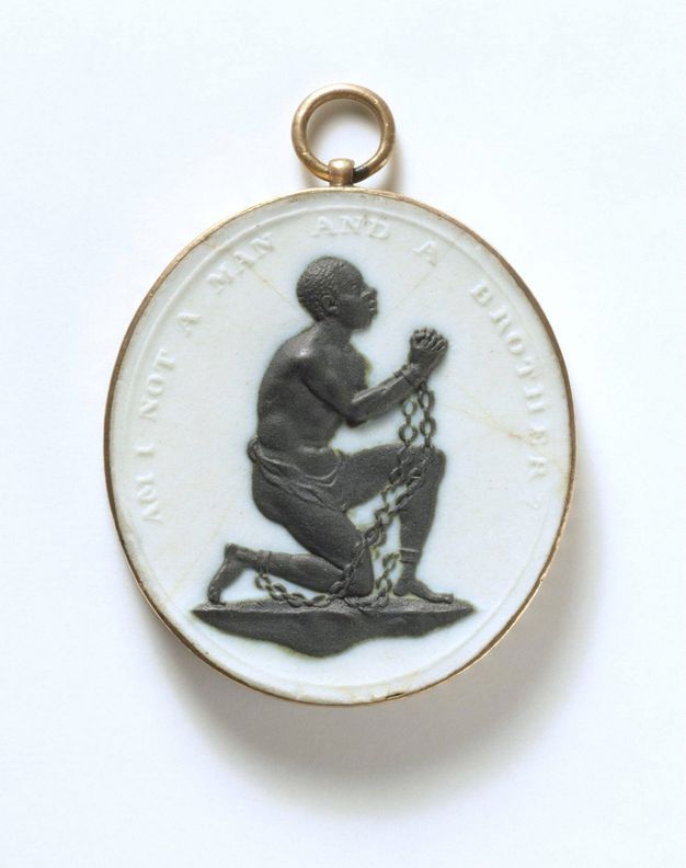 Medallion. In black against a white oval background, a man kneels and lifts his clasped hands. Chains connect shackles on his wrists to more on his ankles. His torso hunches forwards as he looks up. Raised lettering around the perimeter reads, “Am I not a Man and a Brother?” in all caps.
