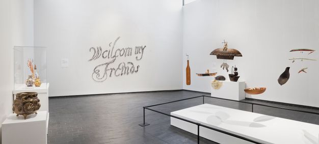 Installation. From one corner we look across a room with a grey stone floor and white walls. Sculptures sit on rectangular white bases, and other works, perhaps mobiles, hang from the ceiling in a group over a low white platform. Writing on the far wall appears to be made with tiny brown dots, and it reads, “Weilcom my Friends”.