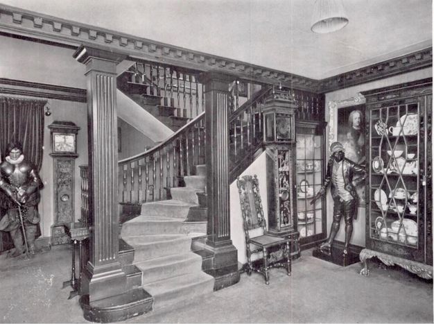 Black and white photograph. A zig-zagging staircase framed by fluted piers creates a corner, in which stand a chair, grandfather clock, two glass-fronted cabinets full of ceramic, and a life-sized, free-standing person. Details are difficult to make out in this image, but the person wears a coat with tails, knee-length breeches and stockings, and a long vest.