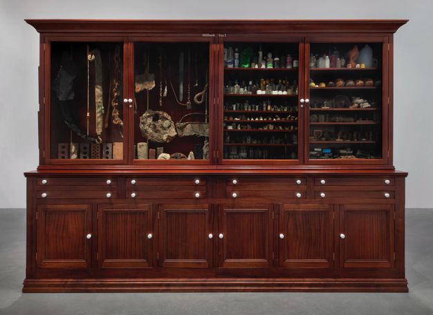 Installation. Bricks, stones, bones, chains, glass bottles, jugs, and other objects fill a glass-fronted wood cabinet with four wide doors.