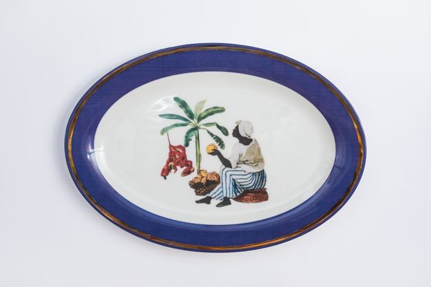 Porcelain. A dark-skinned woman sits near a palm tree holding yellow fruit in one hand at the centre of this white oval plate. She wears a white head covering and shirt, and blue-and-white striped, wide-legged pants. The cobalt-blue rim is edged in gold.