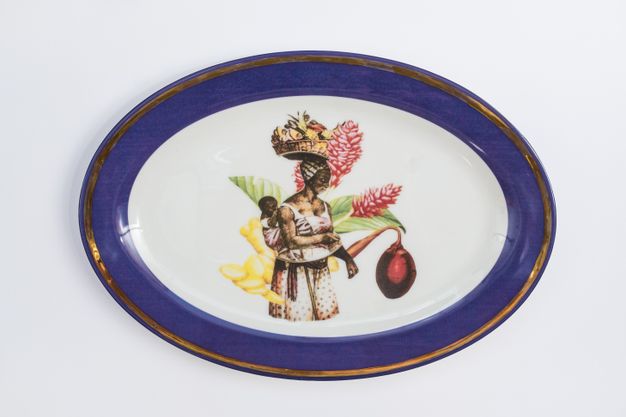 Porcelain. A woman carries a baby on her back at the centre of this white oval plate. Both are dark skinned. The woman balances a basket of fruit on her head and is backed with an oversized flower. The cobalt-blue rim is edged in gold.