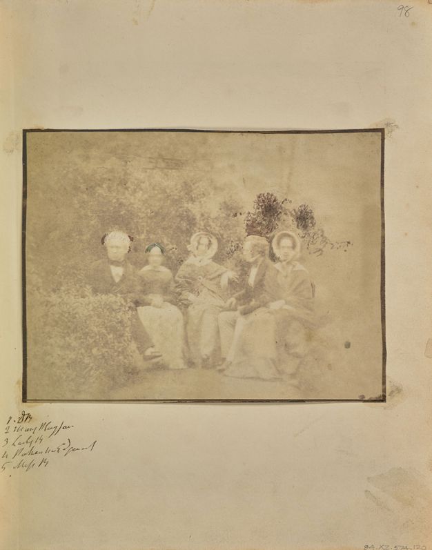 Black and white photograph. Two men and three women sit in a row facing us, in a garden or park setting. The image is pale shades of ash brown, mostly faded with a few notably dark areas.