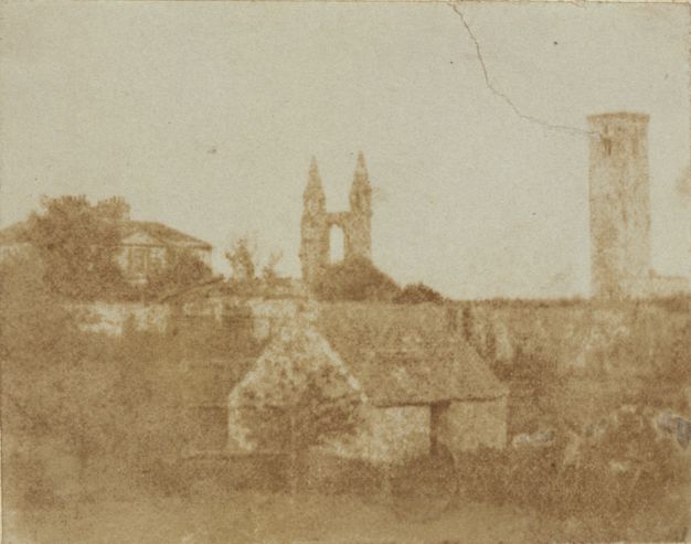 Black and white photograph. In tones of bronze and fawn brown, a small structure sits in front of a wall the same height as its pitched roof. A tower and ruins of a cathedral are outlined against the blank sky in the town beyond the wall.