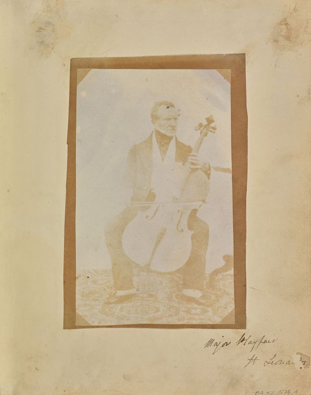 Black and white photograph. In tones of copper brown and tan, a man sits behind a cello, the bow resting on the strings. The other hand is on the neck of the instrument where it meets the body. The pale-skinned, clean-shaven man looks off to our right.