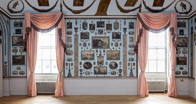 Installation. A pale blue wall has two windows draped with rose pink curtains. Prints are carefully and tidily arranged in the spaces between and around the windows.
