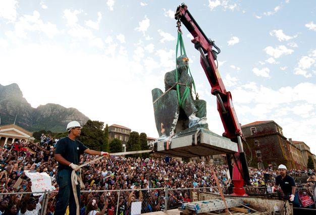 A crane picks up statue of a seated man as a worker wearing a hard hat guides it with a strap. Hundreds of people look on from the background under a blue sky.