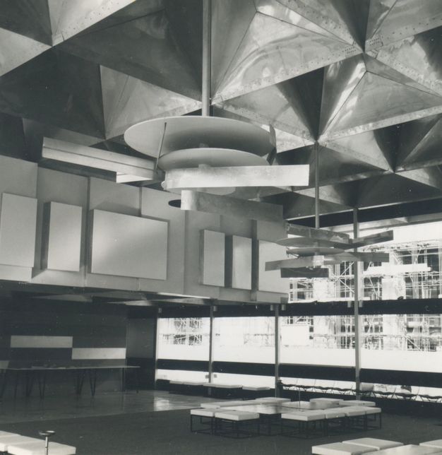 Architecture. Black and white photograph of an interior building filled with abstract designs, from the reflective three-dimensional grid ceiling, to the layered rectangular wall panels, and hanging mobile sculptures of circular plates and rectangular beams.